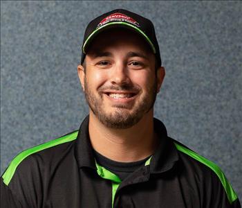 Male employee Ryan poses in a black shirt and hat, with SERVPRO logo, light beard
